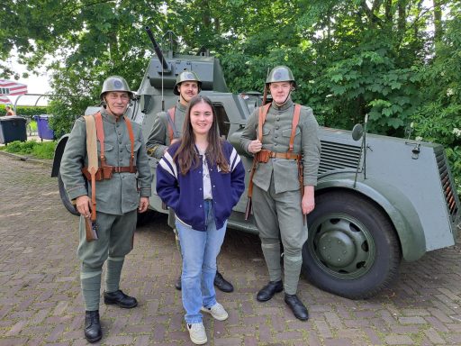 Vrouw tussen drie mannen in historische militaire uniformen, met een voertuig op de achtergrond.