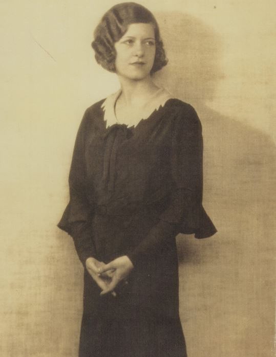 Portret van een vrouw in een zwart jurk met een lichte kraag, staand met gevouwen handen.